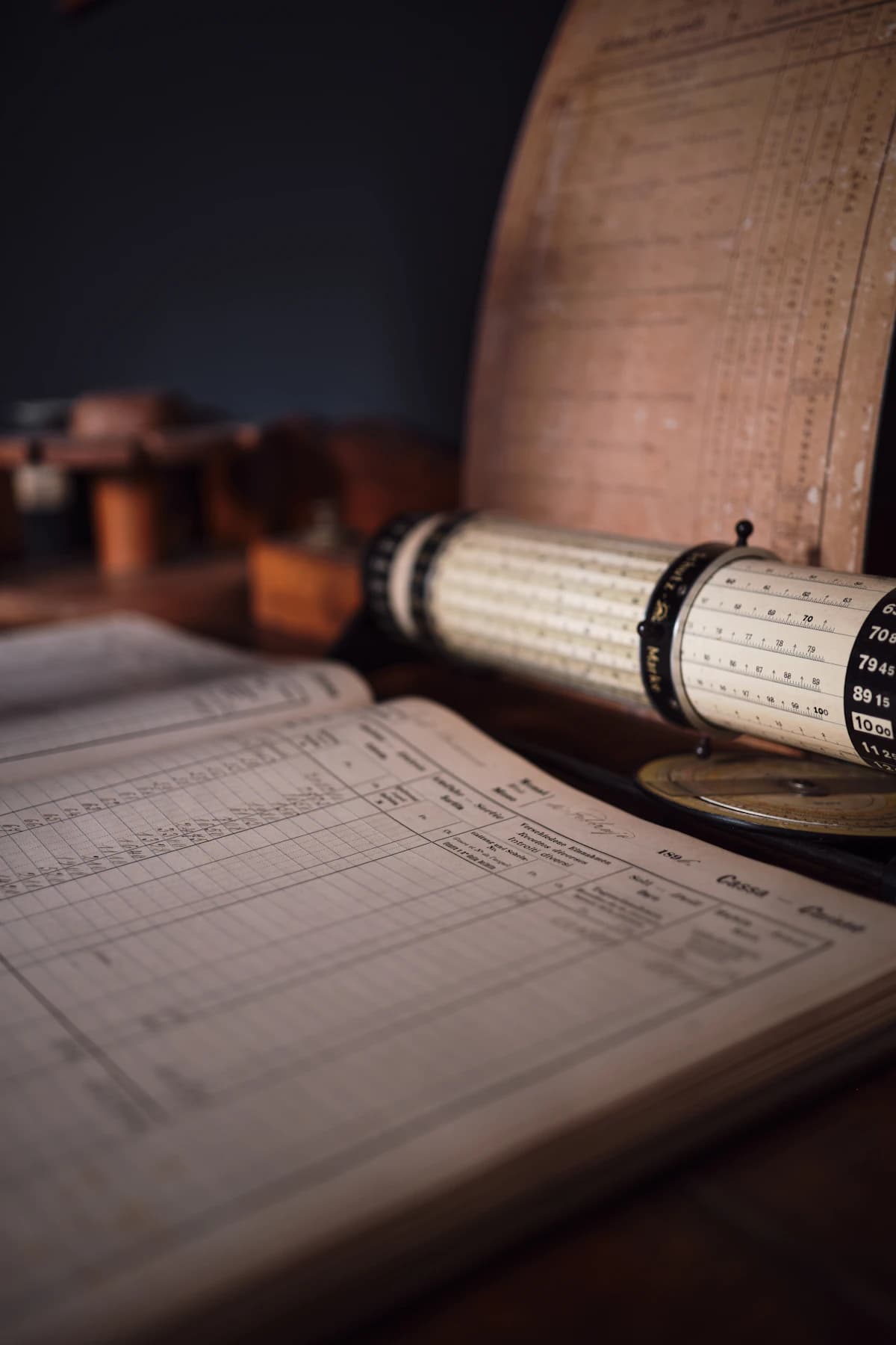 A bound ledger and a brass calculating instrument on a desk, payroll figures worked by hand.